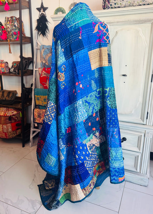A handcrafted silk patchwork quilt with blue as the dominant color, displayed in a room setting with various decorative items in the background.