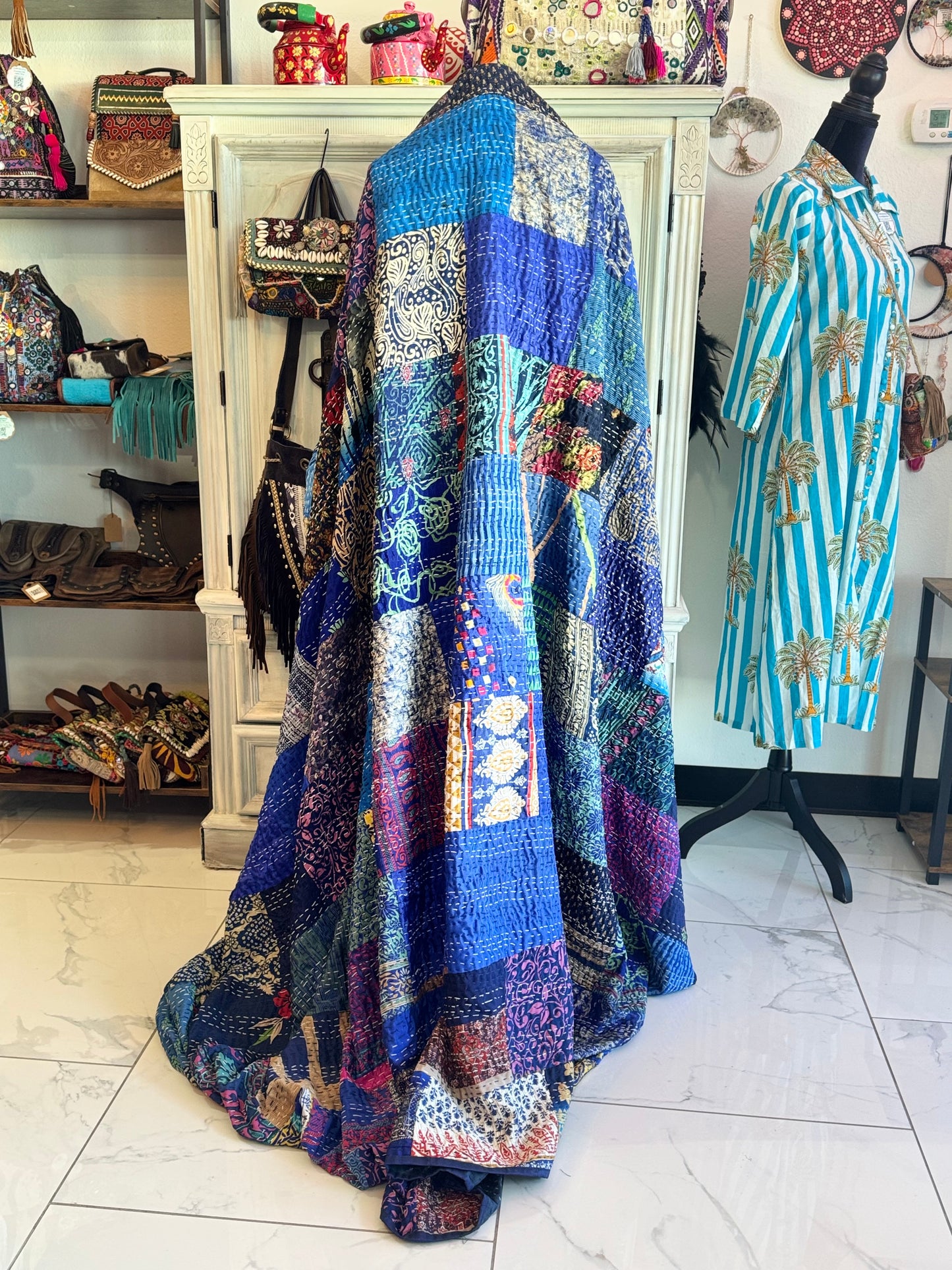 Colorful silk and cotton handmade patchwork kantha quilt draped over a mannequin in a store setting with various items in the background.