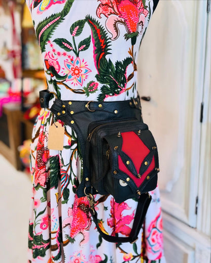 A black leather fanny pack displayed on a mannequin with a floral dress. The fanny pack has adjustable straps and is shown in a front view, highlighting the buckle closures and rivet details.