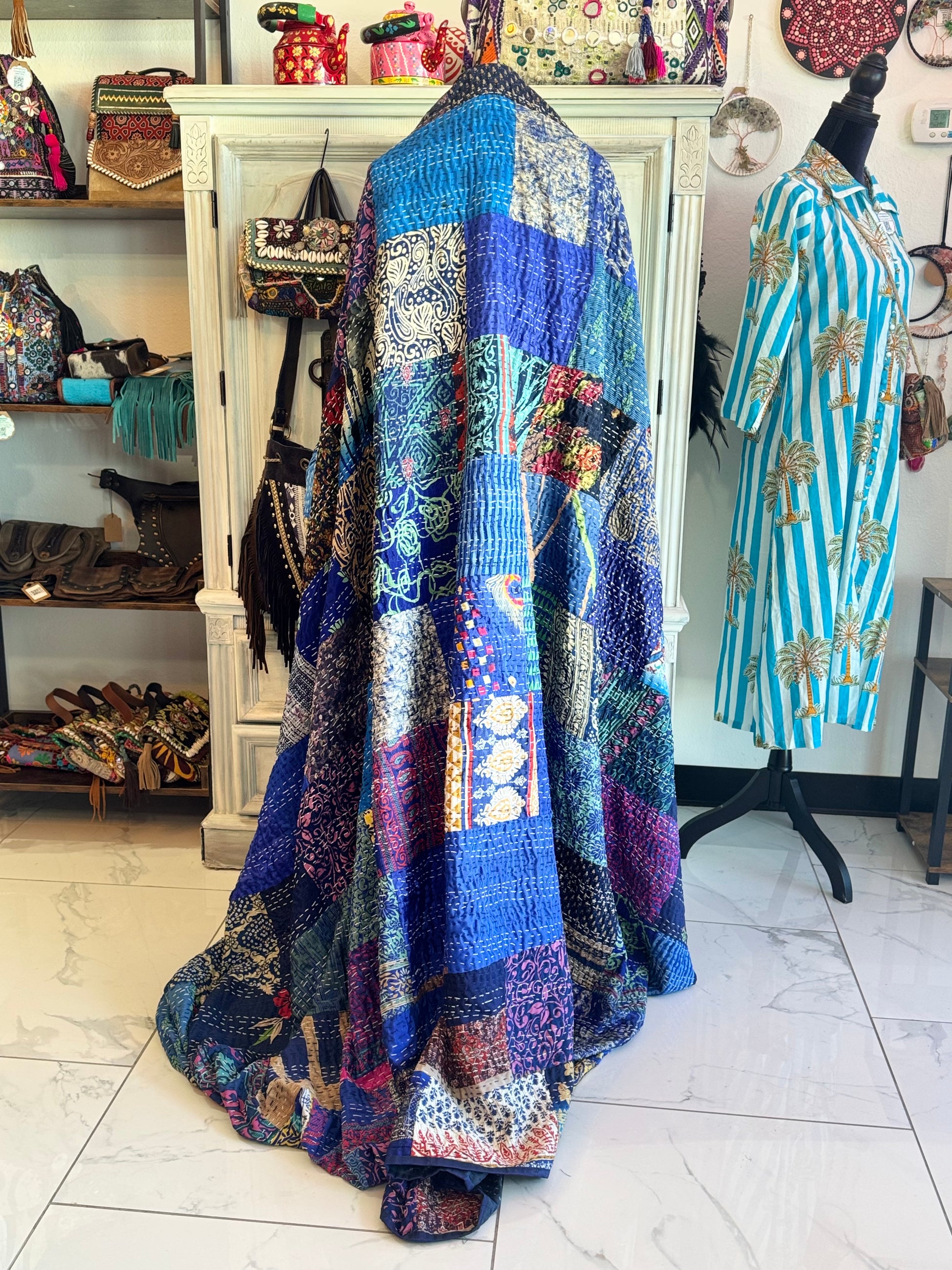 Colorful silk and cotton handmade patchwork kantha quilt draped over a mannequin in a store setting with various items in the background.